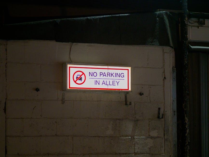 A rectangular illuminated sign mounted on a light-colored brick wall displaying the message 'No Parking in Alley' with a red circle and diagonal line over a car icon. The sign features purple and red text on a white background, bordered by a thin red line. The wall appears to be part of a building exterior, with lighting that suggests evening or night. To the right of the sign, a metal conduit runs vertically along the wall. The setting is relevant to house removals and moving logistics, indicating restricted parking areas on a street or property in Nunhead, where Man with Van Nunhead provides removals services. The sign aids in understanding local parking regulations that affect furniture transport and home relocation processes.