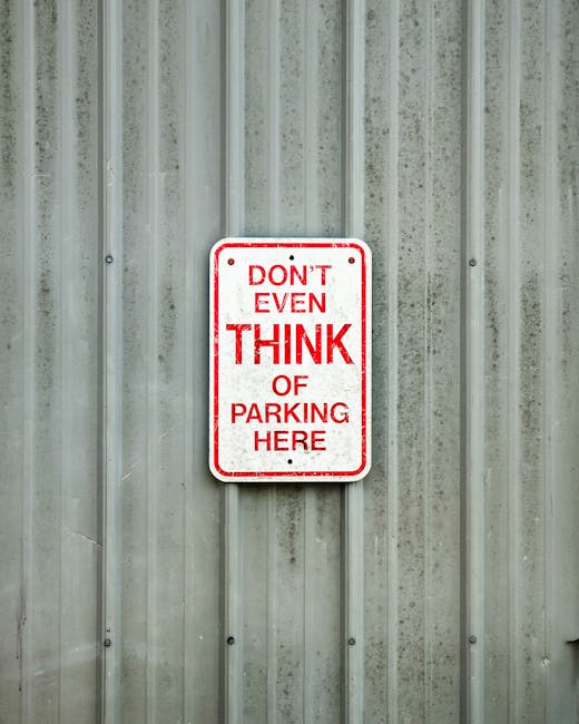 A rectangular metal sign mounted on a vertical, corrugated steel wall with a weathered, slightly dirty appearance. The sign has a white background bordered with a thin red line and displays the message in red capital letters: 'DON'T EVEN THINK OF PARKING HERE.' The background wall has a utilitarian look typical of industrial or service areas. This image relates to parking restrictions in Nunhead, which can impact house removals and moving logistics. The sign's placement suggests a controlled or restricted parking zone, which is relevant for planning local furniture transport and loading during home relocation processes, and the environment indicates an outdoor or loading area for a property or commercial premises, supporting the theme of managing parking rules to avoid fines during moving activities.