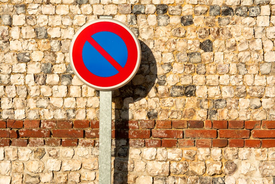 A rectangular illuminated sign mounted on a light-colored brick wall displaying the message 'No Parking in Alley' with a red circle and diagonal line over a car icon. The sign features purple and red text on a white background, bordered by a thin red line. The wall appears to be part of a building exterior, with lighting that suggests evening or night. To the right of the sign, a metal conduit runs vertically along the wall. The setting is relevant to house removals and moving logistics, indicating restricted parking areas on a street or property in Nunhead, where Man with Van Nunhead provides removals services. The sign aids in understanding local parking regulations that affect furniture transport and home relocation processes.