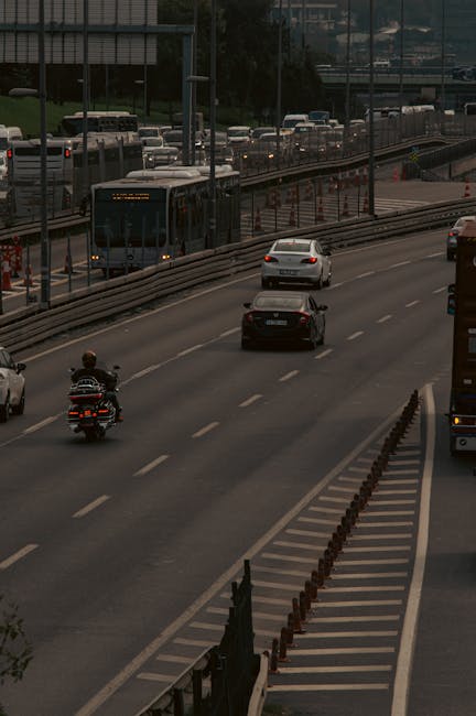 A multi-lane urban road with several vehicles, including a motorcycle, cars, and a bus, moving in various directions. The bus is traveling on the left side of the image, with a digital display indicating it is part of a public transport route. Adjacent to the main lanes, there is a dedicated right-turn lane separated by small orange and black plastic bollards and a striped pedestrian refuge area with paving stones. The roadway is bordered by barriers and street furnishings, with a slight incline visible in the background where additional traffic is present. The scene is captured during daylight with natural lighting, and the environment suggests a busy city area that might be relevant for assessing home relocation routes or transportation logistics in relation to house removals, as possibly discussed on the Man with Van Nunhead website.