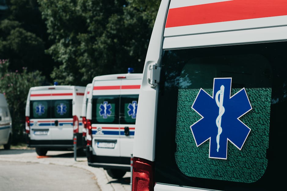 Three parked ambulances with blue medical emblems on their rear doors are situated on a paved area near a green, tree-lined background. The ambulance closest to the camera features a large blue Star of Life symbol with a white rod of Asclepius in the center, displayed on its rear window. The vehicles are positioned in a row, slightly angled to the left, with the one in the foreground showing a side door and part of the rear. The scene is outdoors, illuminated by natural daylight, indicating a temporary emergency vehicle parking area, potentially related to urgent assistance or transport services. This setup may involve logistical aspects of emergency or urgent medical relocation, relevant to services like those provided by Man with Van Nunhead, particularly in contexts requiring rapid transport or logistical management of equipment and personnel during house or property removals or emergency evacuations.