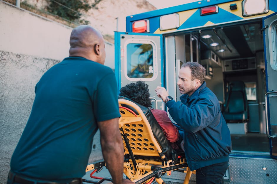 Two men are engaged in a house removal process outdoors during daylight. One man, with short dark hair and wearing a navy jacket, is standing beside an open ambulance or rescue vehicle, actively checking or adjusting a piece of medical or safety equipment. The other man, with a shaved head and dressed in a dark t-shirt, is assisting by holding a stretcher or wheeled chair, which is positioned next to the vehicle. The stretcher has a black fabric covering and an orange plastic frame. Behind them, the vehicle's interior is visible with shelves and compartments likely containing moving or emergency supplies. The scene is set on a paved area with a neutral wall in the background, and there is natural light illuminating the scene clearly. This incident involves the handling of equipment that could be used in emergency or medical transport, aligning with the logistical and safety considerations of home relocations or emergency eviction removals that Man with Van Nunhead may provide, particularly in urgent or time-sensitive situations.