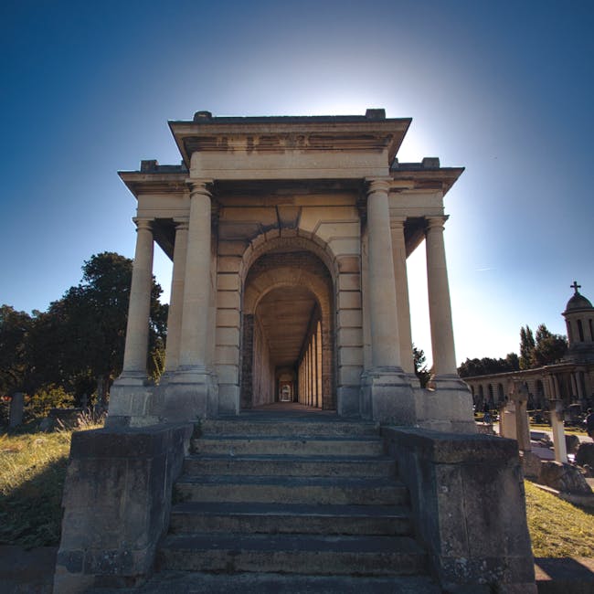 A stone memorial structure with a classical architectural design featuring a set of worn stone steps leading up to an arched passageway supported by four large, round columns. The passage extends through the structure, showing a long corridor that appears to be part of a larger building or monument within a cemetery grounds, with trees and additional structures visible in the background. The area around the steps is outdoors with clear weather and bright sunlight, casting shadows on the stone surfaces, as part of a scene that could be associated with historic site visits or arrangements for house removals involving careful transport of belongings near heritage landmarks. Man with Van Nunhead’s removals service may utilize such sites for documentation or staging tasks related to home relocation and furniture transport, ensuring a professional and secure moving process.