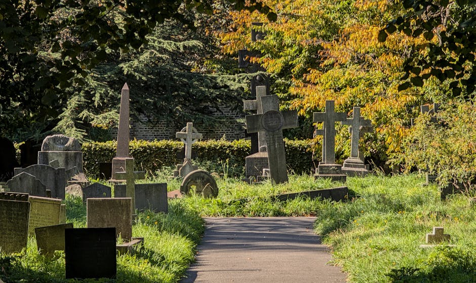 A pathway in Nunhead Cemetery lined with various gravestones and memorial crosses situated on either side of the paved walkway, surrounded by lush green grass and trees with autumn foliage in shades of yellow and orange. In the background, dense foliage provides a natural canopy, with some of the gravestones leaning or partially obscured by vegetation. The scene is illuminated by natural daylight, highlighting the somber and tranquil atmosphere. In a moving or relocation context, this setting aligns with the process of home relocation near historic cemeteries, where [COMPANY_NAME] may assist with furniture transport, packing, and loading to ensure respectful handling of belongings within such environments.