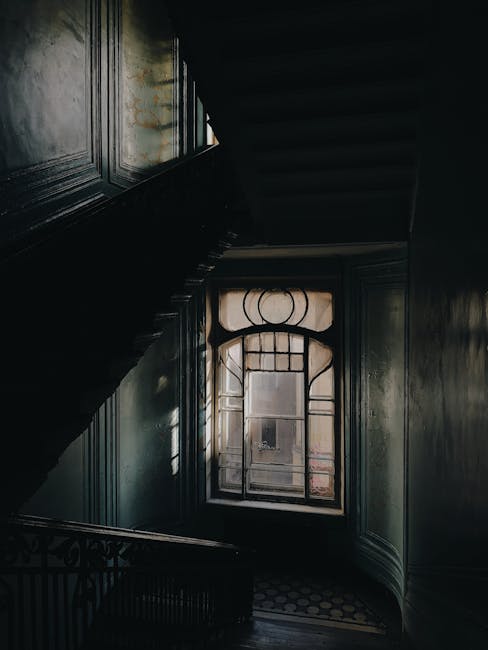 The image shows the interior of a narrow Victorian house staircase in Nunhead, featuring wooden paneling on the walls and a dark staircase. A large window with decorative wrought iron design is visible at the landing, allowing natural light to illuminate the space. The window reveals a view of the exterior brick wall of the building. The staircase appears steep and confined, emphasizing the challenges of navigating such narrow staircases during home relocation processes. This setting likely belongs to an older property in Nunhead, requiring careful handling during furniture transport and packing within the restricted space. The image is taken from the lower landing, looking upward toward the window, with shadows cast across the walls and stair treads, illustrating the environment that specialists like Man with Van Nunhead manage when handling house removals in historic, narrow staircases.