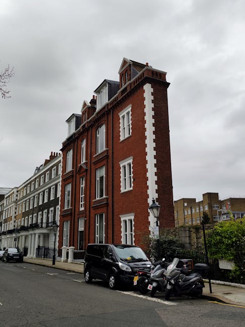 Photograph of a narrow, multi-storey Victorian house in Nunhead constructed of red brick with white decorative accents around the windows and a distinctive corner turret. The building has large, vertically aligned windows on each floor, some with white frames and sash windows. It is situated on a street with other similarly styled houses, featuring mixed-use facades in white and grey tones. In front of the house, a black van and several scooters are parked on the pavement, indicating recent or ongoing house removal activities. The scene is captured during overcast weather, with grey clouds visible in the sky, and the street appears quiet, typical of residential areas. This image was taken as part of a house relocation effort by Man with Van Nunhead, highlighting the logistics involved in moving furniture and household items through narrow, urban environments during a packing and moving process.