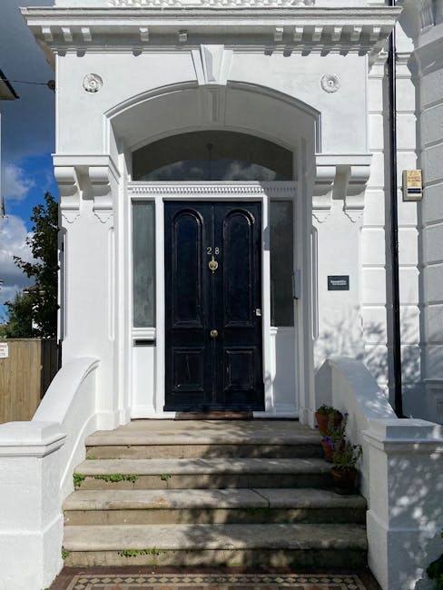 The image shows the interior of a narrow Victorian house staircase in Nunhead, featuring wooden paneling on the walls and a dark staircase. A large window with decorative wrought iron design is visible at the landing, allowing natural light to illuminate the space. The window reveals a view of the exterior brick wall of the building. The staircase appears steep and confined, emphasizing the challenges of navigating such narrow staircases during home relocation processes. This setting likely belongs to an older property in Nunhead, requiring careful handling during furniture transport and packing within the restricted space. The image is taken from the lower landing, looking upward toward the window, with shadows cast across the walls and stair treads, illustrating the environment that specialists like Man with Van Nunhead manage when handling house removals in historic, narrow staircases.
