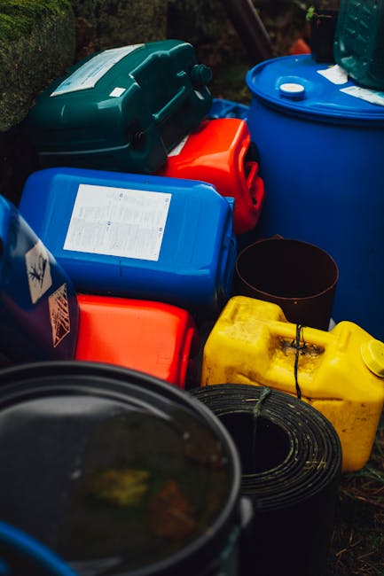 A collection of brightly coloured plastic fuel cans, including blue, yellow, and red containers, arranged on the ground outside a property. The cans vary in size, with some featuring handles and spouts, while others are rectangular with labels indicating contents. There are also coiled black hoses or cables placed among the fuel cans. The containers are positioned on a paved or gravel surface, with a backdrop of greenery, and appear to be awaiting transportation or disposal as part of a house removal or home relocation process. The scene is well-lit, highlighting the vibrant colours of the fuel containers and the details of the packaging materials, such as plastic wrap on some items. This image depicts the handling and organization of hazardous or bulky waste materials, which are typical considerations during professional house removals and moving services provided by [COMPANY_NAME], especially in the context of adhering to waste management rules for removals in Nunhead.