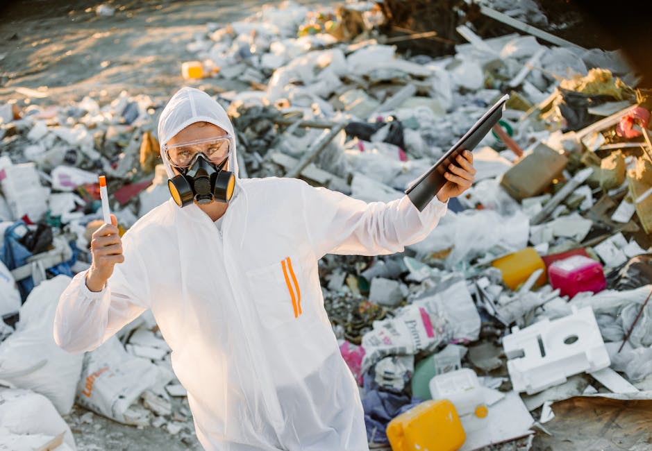 A person wearing protective clothing, including a white coverall, goggles, and a respirator mask, stands amid a large pile of mixed waste materials, including plastic containers, cardboard boxes, and packaging debris, at what appears to be a cleanup or waste disposal site. The individual holds an orange litter picker in one hand and a tablet device in the other, seemingly assessing or documenting the waste. The scene is outdoors, with sunlight illuminating the rubbish and the person, indicating a waste management or environmental cleanup operation. This image reflects the proper handling and sorting of waste materials, relevant to house removals and the disposal of hazardous or bulky waste, which Man with Van Nunhead can assist with as part of their removals and waste removal services.
