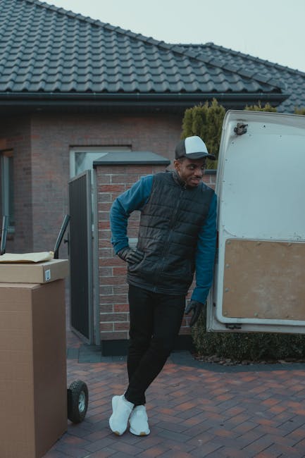 A young male mover with shoulder-length hair, wearing a dark headband and a navy blue shirt, is sitting in the driver's seat of a white moving van. He is smiling and giving a thumbs-up gesture towards