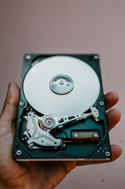 A close-up of a person's hand holding an open hard disk drive (HDD), showing the internal components such as the silver magnetic platter, read/write arm, actuator, and circuit board. The background is
