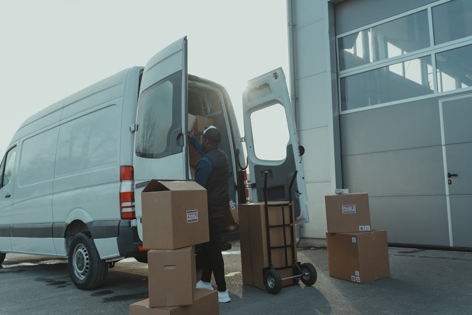 A professional mover from Man with Van Nunhead is loading cardboard boxes and packaging materials into a large white van outside a modern building with glass windows in the background. The mover, wearing a dark vest and cap, is seen placing a cardboard box near the open rear doors of the van, which is situated on a paved surface adjacent to the building. Several other cardboard boxes, some sealed and others unsealed, are arranged on the ground and a hand truck or dolly is positioned nearby, ready for transport. The lighting suggests daylight, possibly during late afternoon, with the sun casting a bright glow behind the building. This scene captures a home relocation or furniture transport process involving packing and loading, reflecting the logistical aspects of house removals in preparation for a move in Southwark, as indicated by the proximity to a parking or loading zone. The image illustrates elements of moving services and the careful handling of household items during a professional home move, consistent with house removal practices aligned with Southwark council permits in SE15, Nunhead.