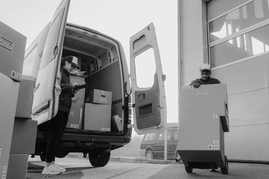 Two movers from Man with Van Nunhead are loading cardboard boxes into the back of a white moving van involved in home relocation. The moving van has its rear doors fully open, revealing several sealed boxes placed inside, some stacked on top of others. One mover, wearing a cap and casual clothing, stands inside the van checking or organizing items, while another, wearing a safety helmet, is positioned outside the van pushing a large cardboard box on a wheeled trolley. The boxes are made of sturdy cardboard and are labeled with handling symbols. The scene takes place outdoors near a modern building with large glass windows, and additional boxes are visible on the pavement next to the loading area. The lighting is natural, and the environment reflects a typical furniture transport and packing process during a professional move managed by [COMPANY_NAME], supporting efficient house removals and logistical operations.