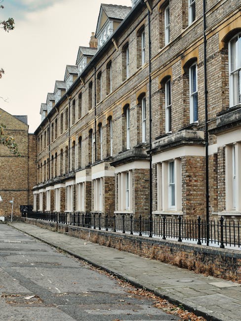A quiet residential street near Nunhead with a narrow asphalt road flanked by brick terraced houses on the left and a paved sidewalk lined with dense green hedge on the right. The houses feature white
