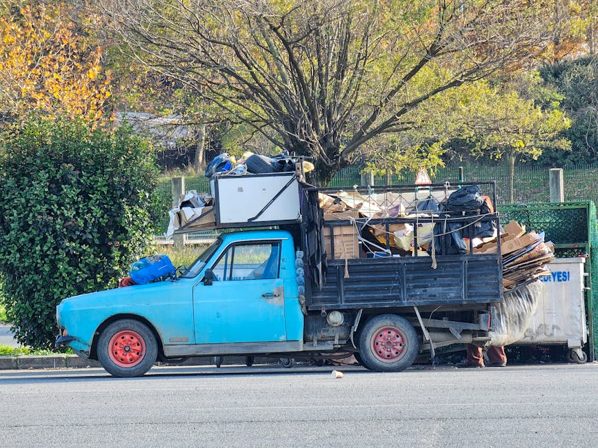 Side view of a small, blue, vintage-style flatbed truck parked on the street during daytime, loaded with various packing materials and household items for home relocation. The vehicle's cab has a slightly weathered appearance, with visible dirt on the body and red-painted wheels. On the flatbed, there are large cardboard boxes, wrapped furniture, plastic containers, and bundled packing materials secured with ropes and straps. Some items are covered with protective blankets, suggesting careful handling during furniture transport and moving logistics. Behind the truck, a tree with green and autumn-colored leaves is visible, indicating an outdoor setting in a suburban area. The truck belongs to Man with Van Nunhead, a removals service specializing in packing and moving. The scene captures the loading process, with objects positioned on the pavement and inside the vehicle, ready to be driven to a new property.
