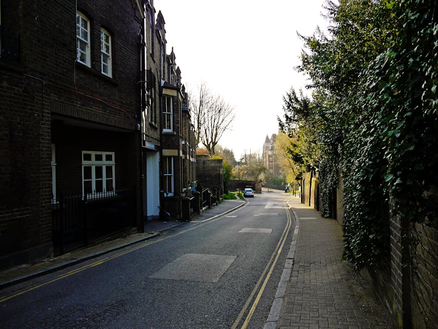A quiet residential street with a narrow paved road and double yellow lines along the curb, flanked by brick terraced houses on the left side featuring bay windows and small front gardens. On the right, there is a sidewalk bordered by a tall, dense hedge and a brick wall. The street is illuminated by soft daylight, with some cars parked in the distance. In the foreground, a moving van from Man with Van Nunhead is parked partially on the pavement, with the rear doors open. Inside the van, there are several cardboard boxes, some covered with plastic wrapping and bubble wrap, along with a large wooden furniture piece and various packing materials. A set of hand trucks or dollies is visible, used for loading and transporting household items during a home relocation. Nearby, a person, partially visible, is carrying a small box towards the van, highlighting the ongoing furniture transport and packing process involved in the moving services for Nunhead Lane and The Old Nun's Head, NUNHEAD.