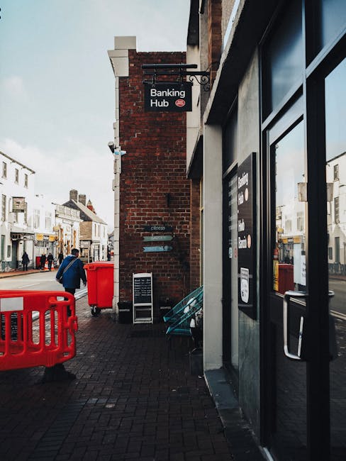 A street scene outside a building with a sign indicating it is a banking hub, showing a brick exterior wall and large glass windows. In the foreground, there is a partially visible black door with a handle, likely the entrance to the banking branch. To the left, a person wearing a blue jacket and carrying a backpack is walking along the pavement. Nearby, a red plastic barrier and a red waste bin are positioned on the sidewalk. Further down the street, additional pedestrians are visible, along with shops and residential buildings with pitched roofs in the background. The street is lined with a mix of local businesses and residential properties, with overcast lighting creating a muted atmosphere. This scene could relate to house removals or relocation services, as the environment includes urban elements such as pathways, signage, and pedestrian activity, and may depict a location where furniture or boxes are being moved or accessed for a home relocation process. Man with Van Nunhead provides services in similar urban settings, supporting smooth logistics for moves within such areas.