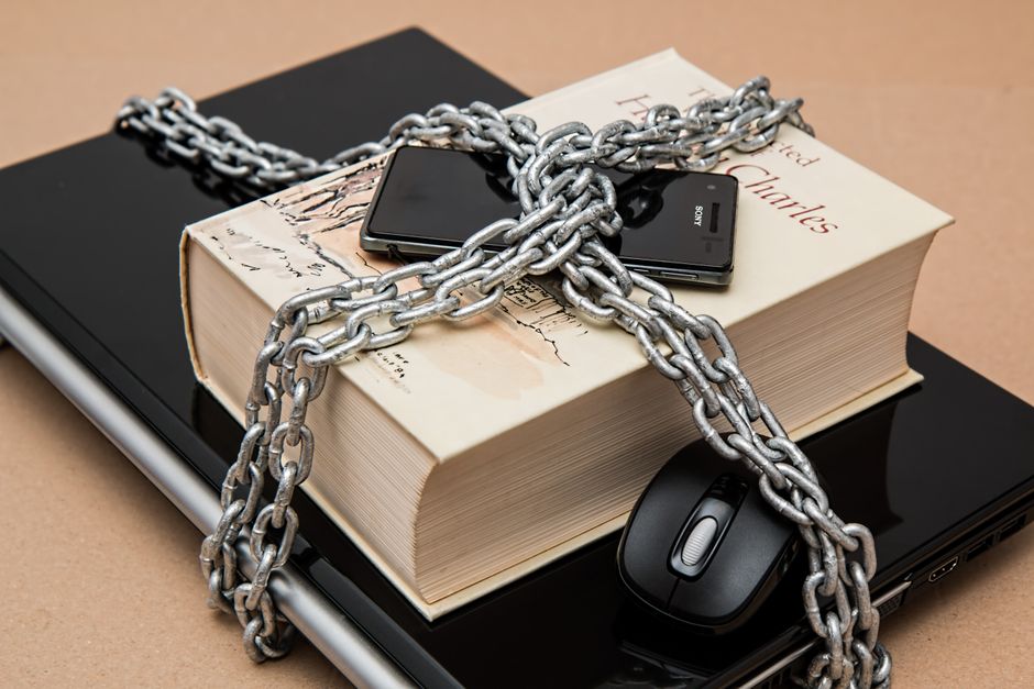 A close-up image showing a large paperback book placed on a closed black laptop, with a black smartphone resting on top of the book. A thick metal chains is draped over the book and devices, crossing in an X-shape. To the right of the book, there is a black computer mouse. The background includes a cardboard surface, and the scene appears to illustrate packing materials and items typically involved in home relocation or furniture transport, with the chains symbolising securing or safeguarding possessions during a move. The setup suggests a preparation stage in a house removals process, highlighting the importance of careful packing and securing of belongings prior to transport. This visual aligns with house removal services offered by Man with Vann Nunhead, focusing on packing, moving, and transportation considerations.